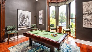A pool table in a room with dark walls, large windows, and green upholstered chairs.