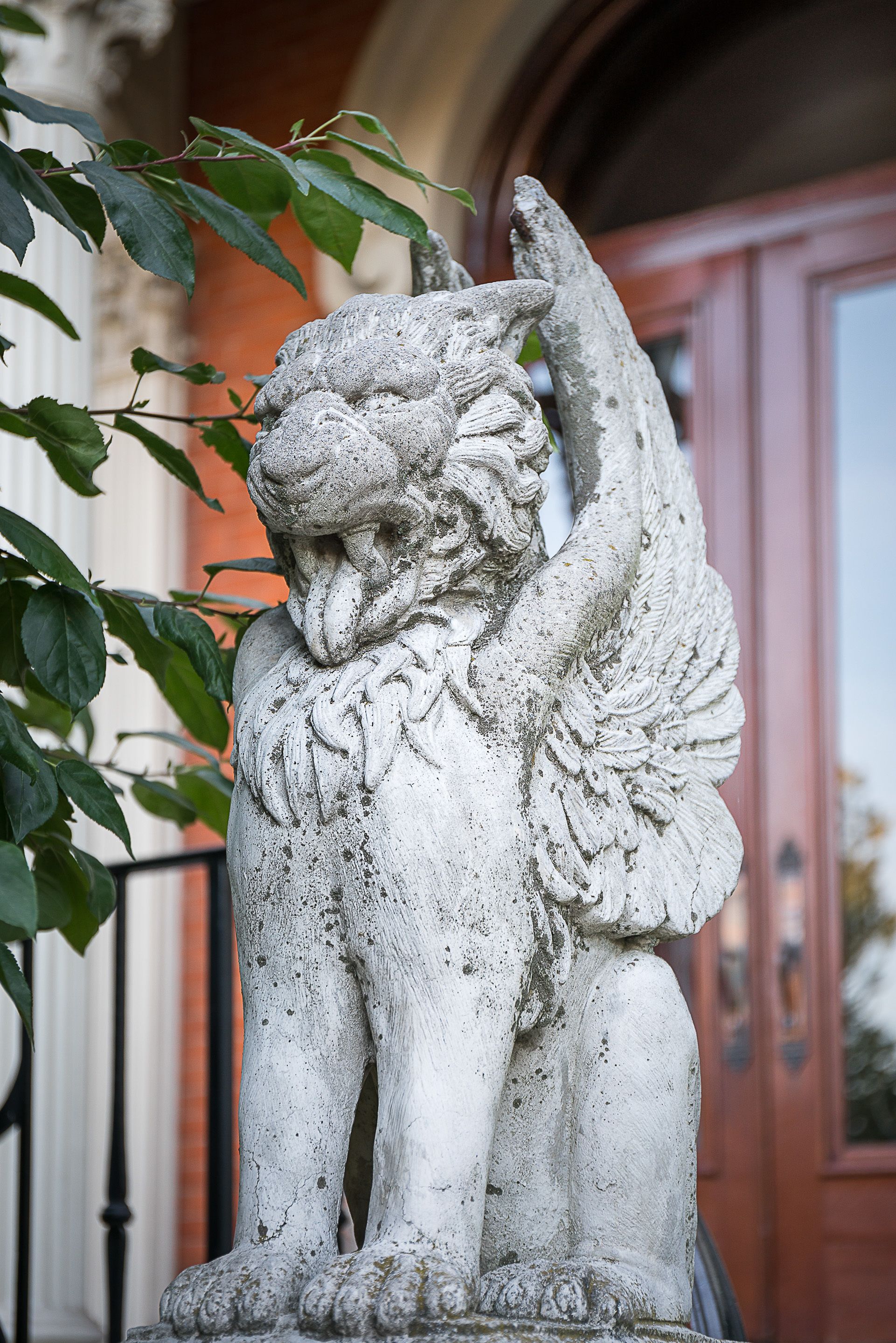 Una estatua de piedra de un león con una hoja en la boca, ubicada frente a la entrada de un edificio con puerta roja y reja de metal.