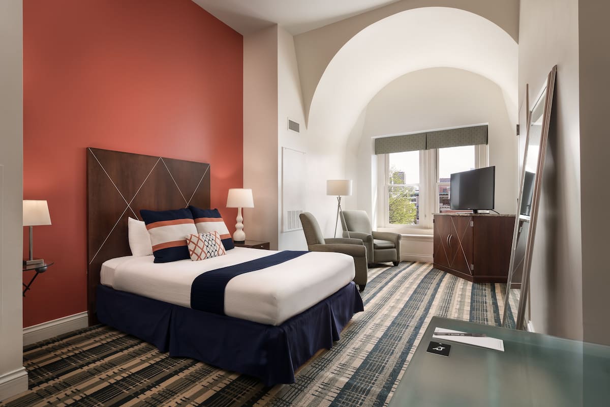 A hotel room with a large bed, two chairs, a desk, a TV, and a window with a view.