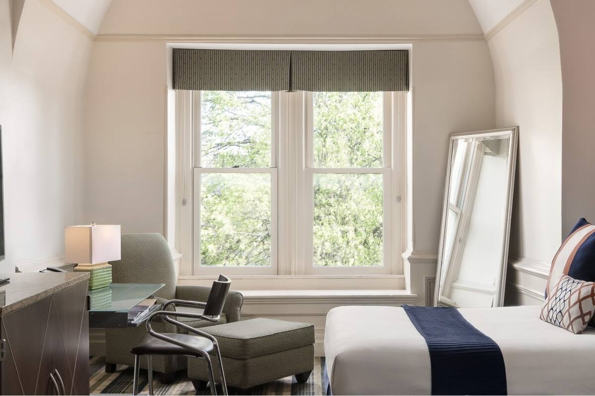 A hotel room with a bed, a desk with a chair, a television, and a window with a view of trees.