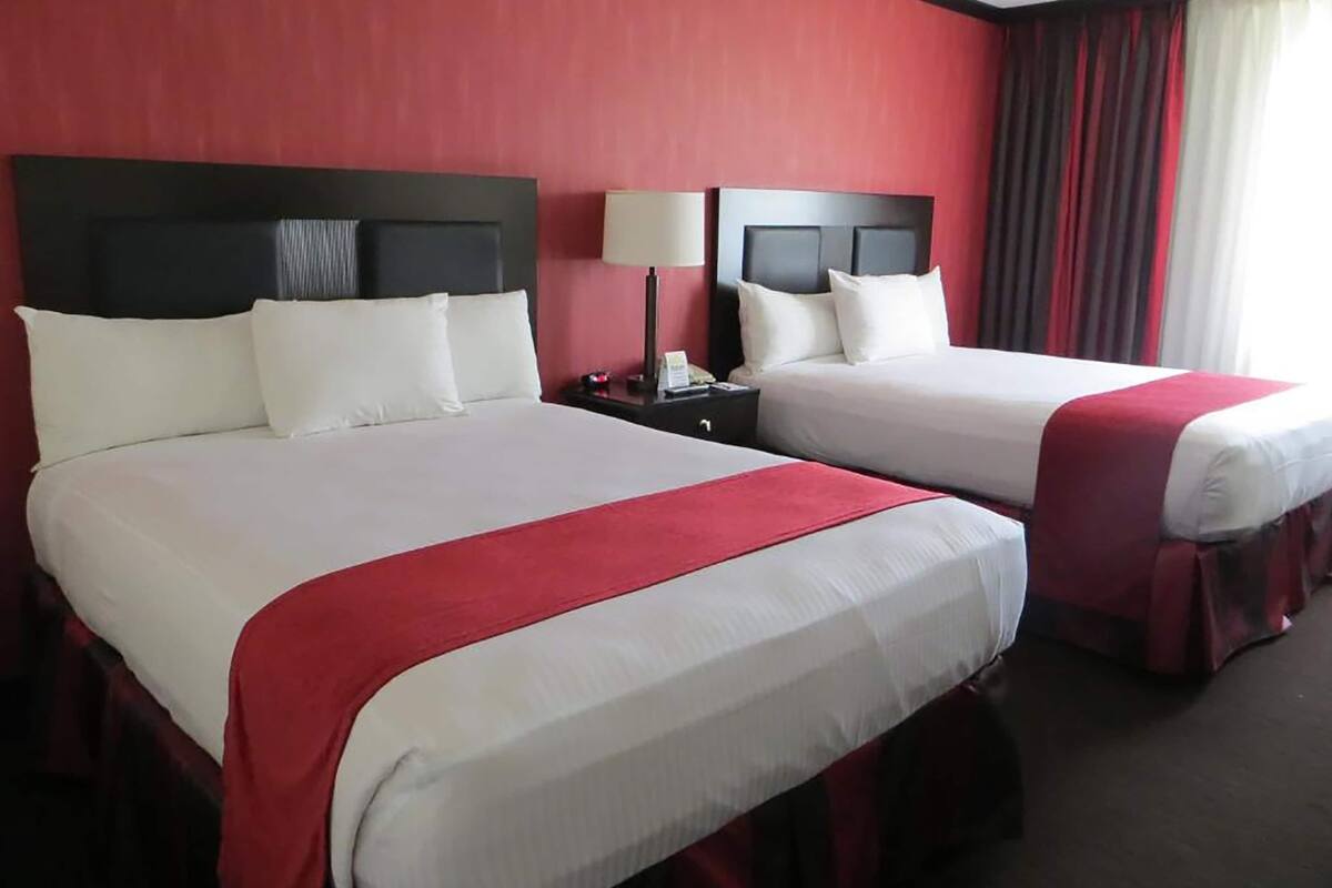 Two adjacent hotel beds with white linens and red accents, a nightstand with a lamp, and a red curtain.