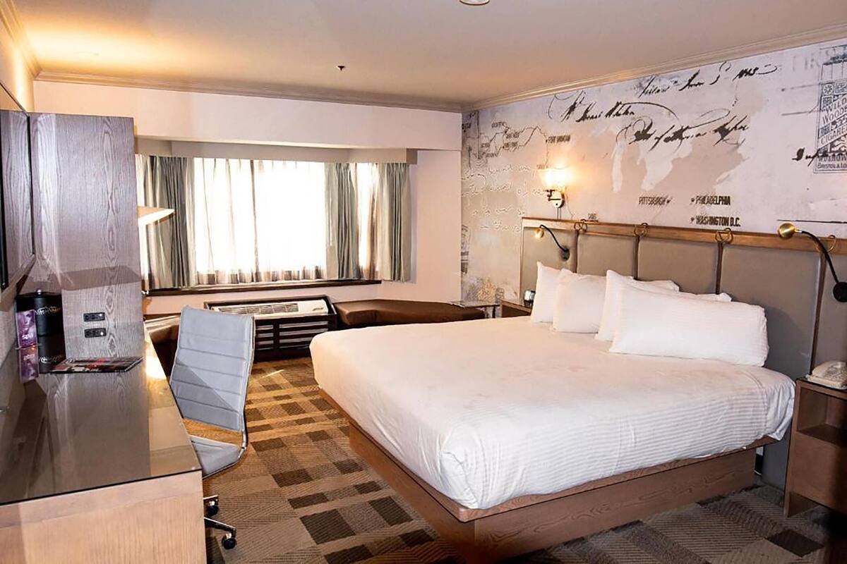 A hotel room with a large bed, a desk with a chair, a map wall, and a window with curtains.