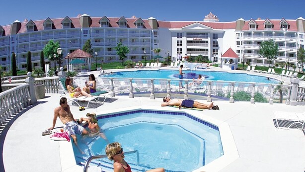 A resort with a large swimming pool surrounded by lounge chairs and a multi-story building with balconies.