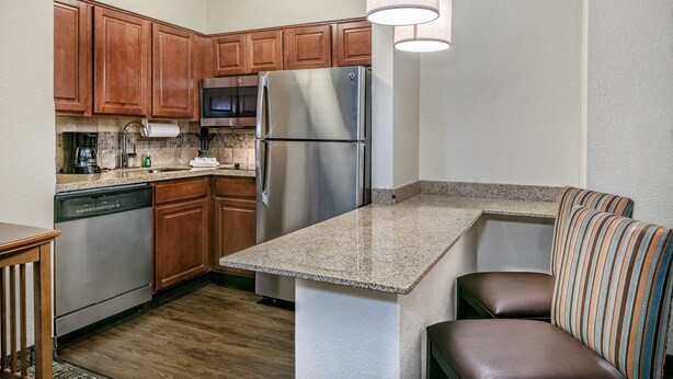 A compact kitchen with wooden cabinets, a stainless steel refrigerator, a dishwasher, and a built-in bench with striped cushions. - Staybridge Suites Round Rock by IHG (Round Rock)