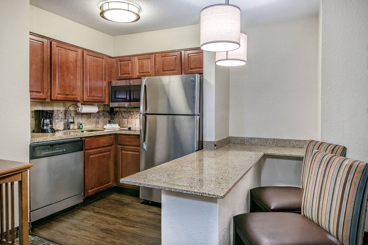 A compact kitchen with wooden cabinets, a stainless steel refrigerator, a dishwasher, and a built-in bench with striped cushions.