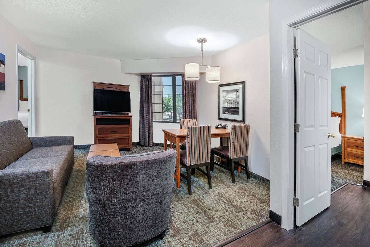 A hotel room with a sofa, armchair, dining table with chairs, a TV, a framed picture, and a door.