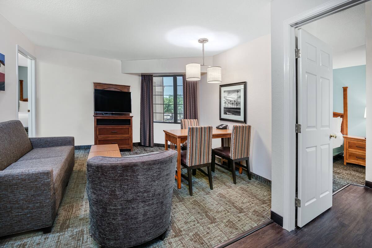 A hotel room with a gray sofa, a round armchair, a dining table with chairs, a flat-screen TV, and a window with curtains.