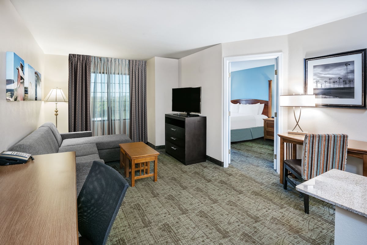 A hotel room with a sofa, a TV, a small wooden table, a desk, and a chair. There is a bed in the background.