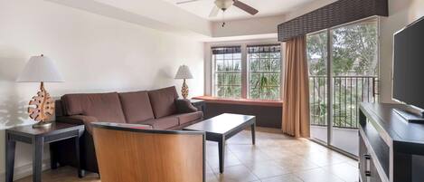 Una sala de estar moderna con un sofá, una mesa de centro y un mueble para la televisión. Tiene un ventilador de techo, ventanas amplias con cortinas y un balcón con vistas.