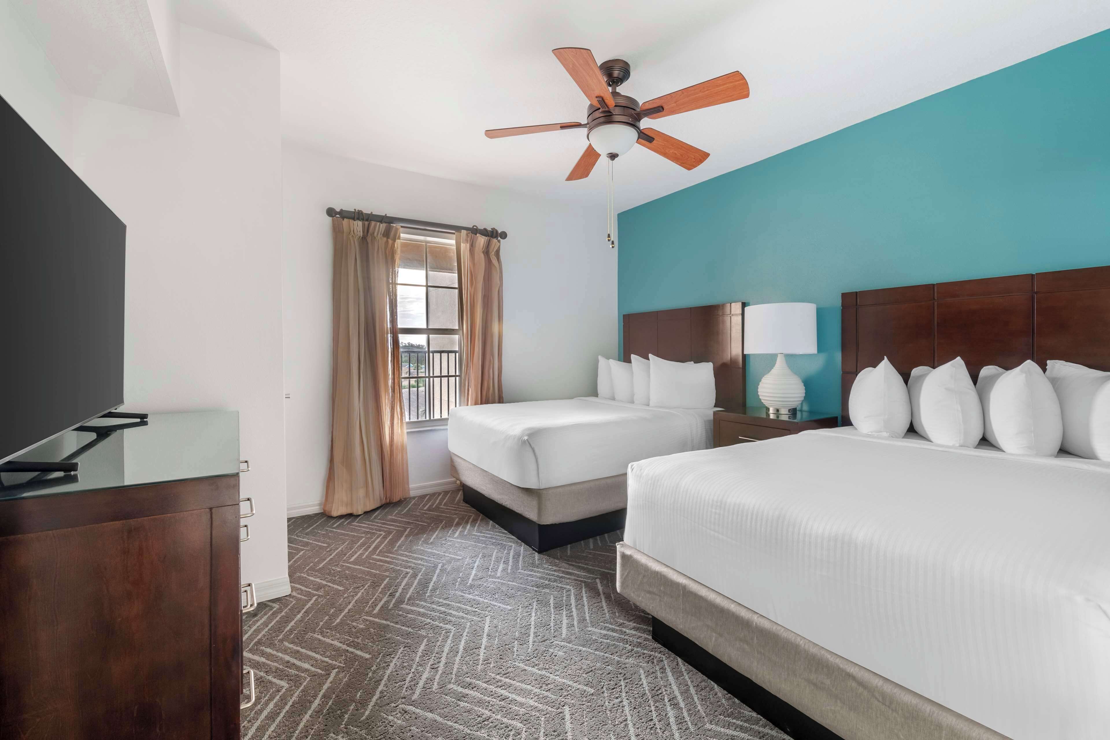 Une chambre d’hôtel avec un lit, un bureau, un ventilateur de plafond et une fenêtre avec des rideaux.