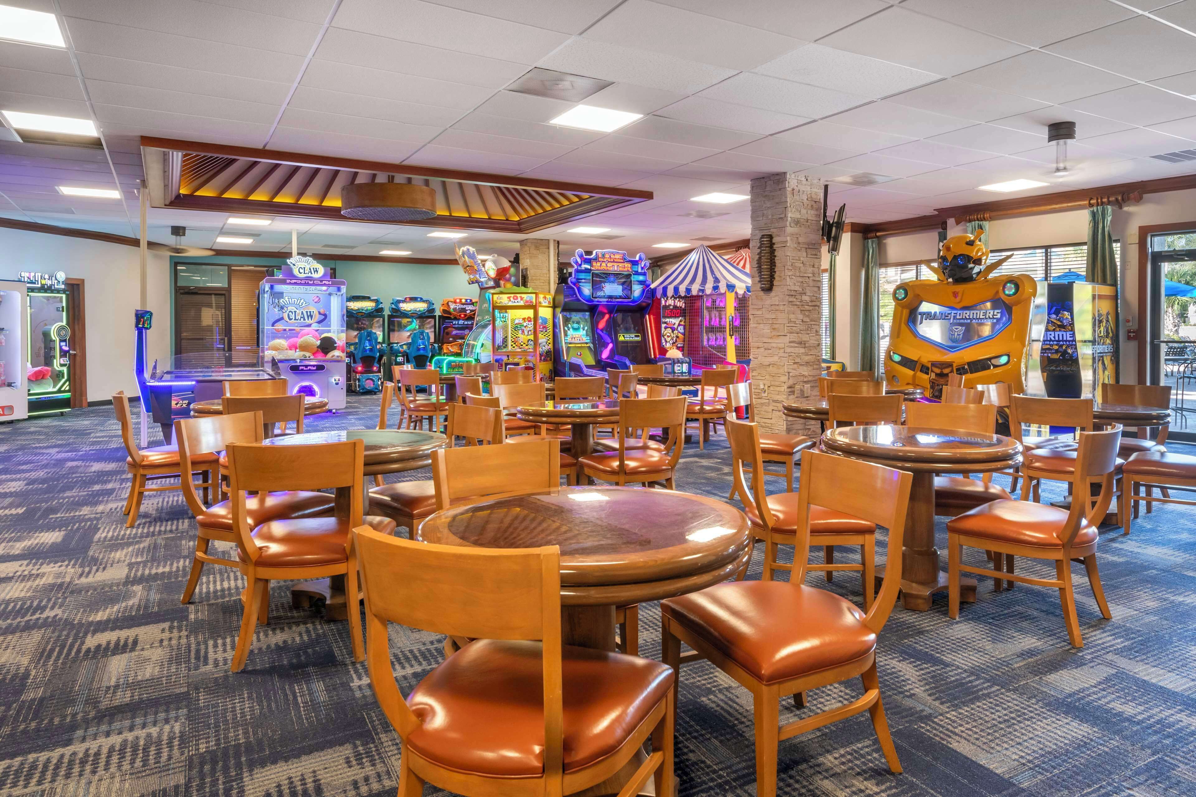 A gaming arcade area with multiple claw machines, a Transformers-themed robot, and several wooden tables and chairs.