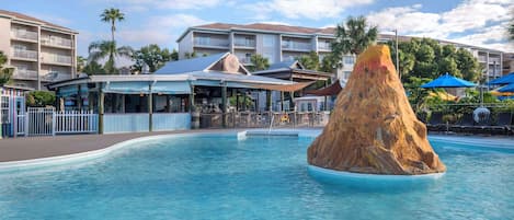 A swimming pool with a rock feature, a covered seating area, and a building with balconies in the background.