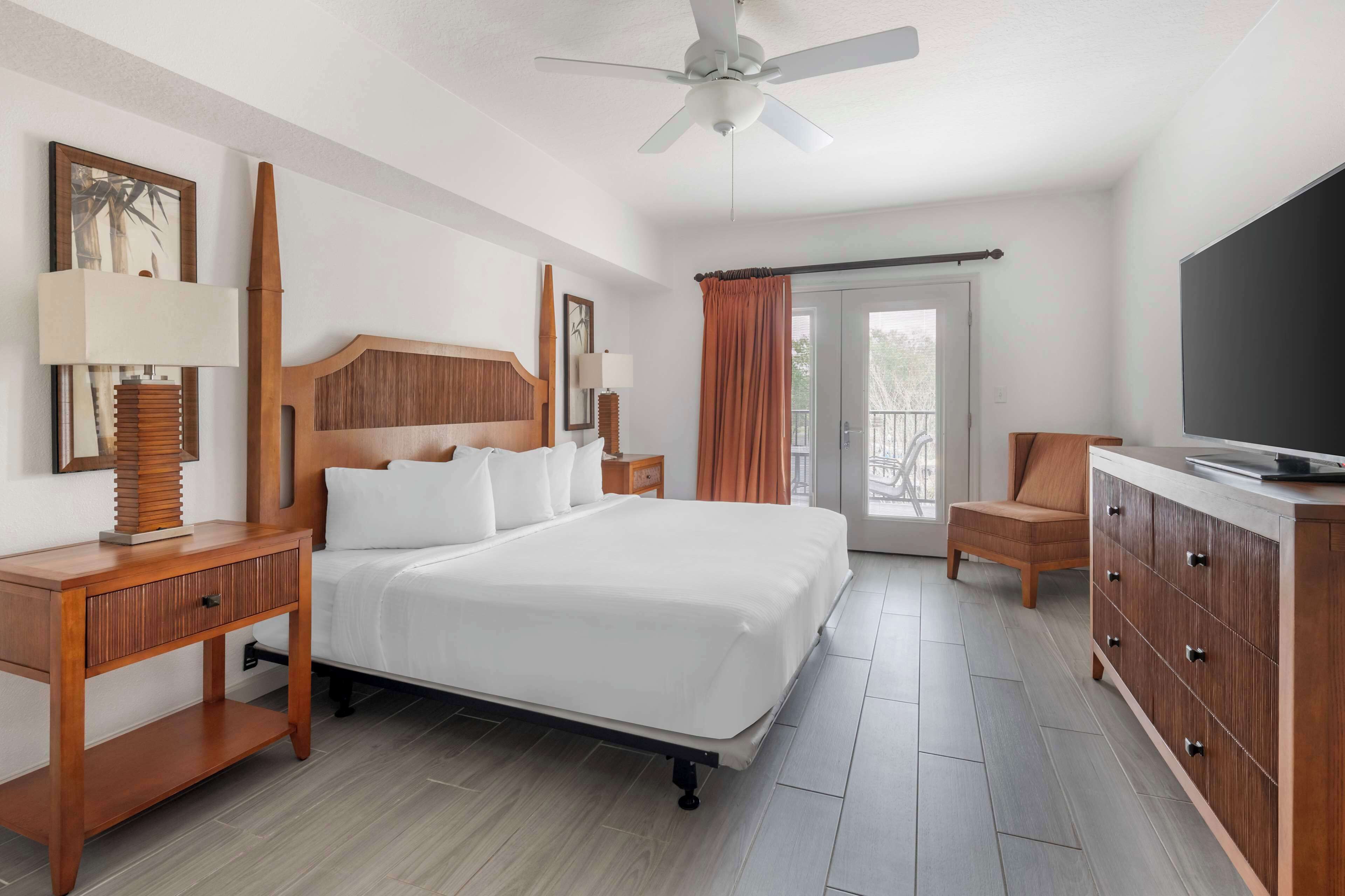 Habitación de hotel con cama, mesitas de noche de madera, televisor y balcón con cortinas.