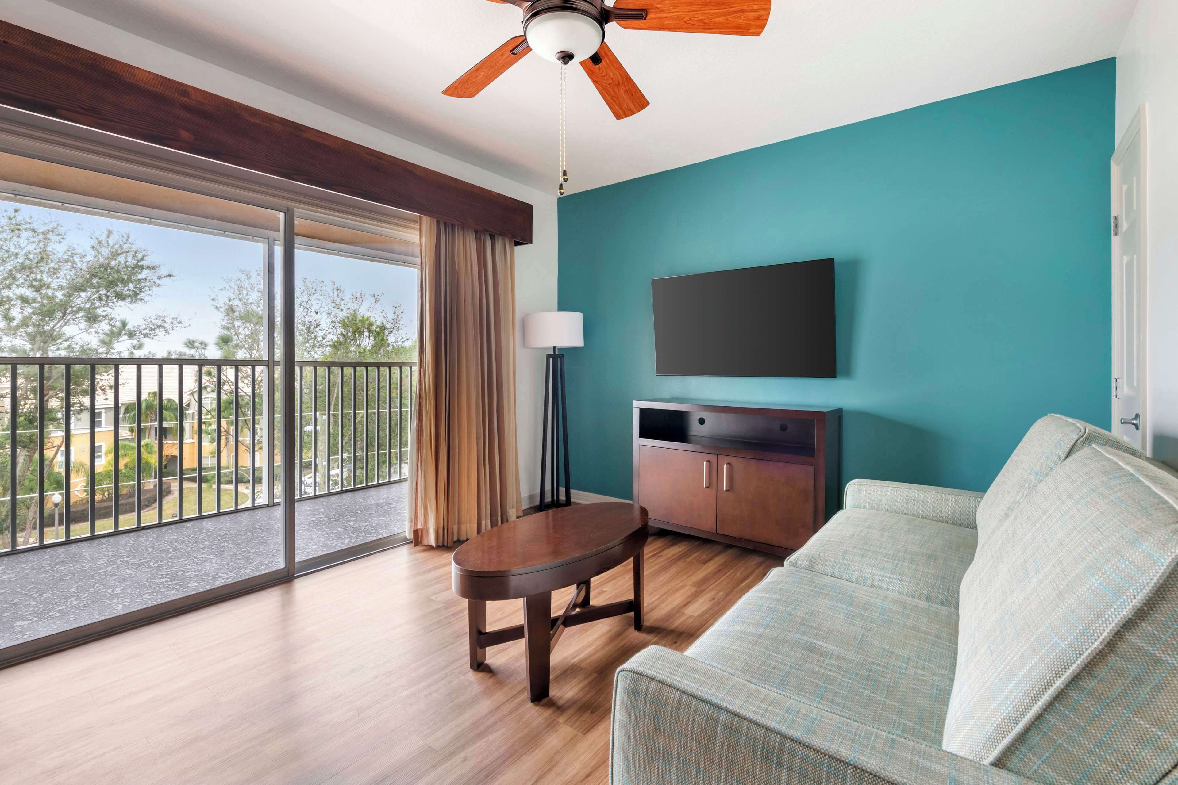 A room with a wooden floor, a teal wall, a flat-screen TV, a sofa, a table, a ceiling fan, and a balcony with a view of the outdoors.