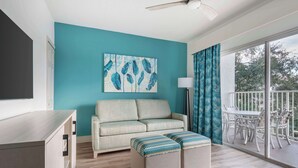 Una sala moderna con una pared de acento color turquesa, un sofá, otomanas, una zona de comedor con sillas blancas y un balcón con vistas.