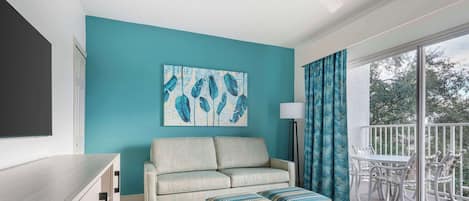 A modern living room with a teal accent wall, a sofa, ottomans, a dining area with white chairs, and a balcony with a view.