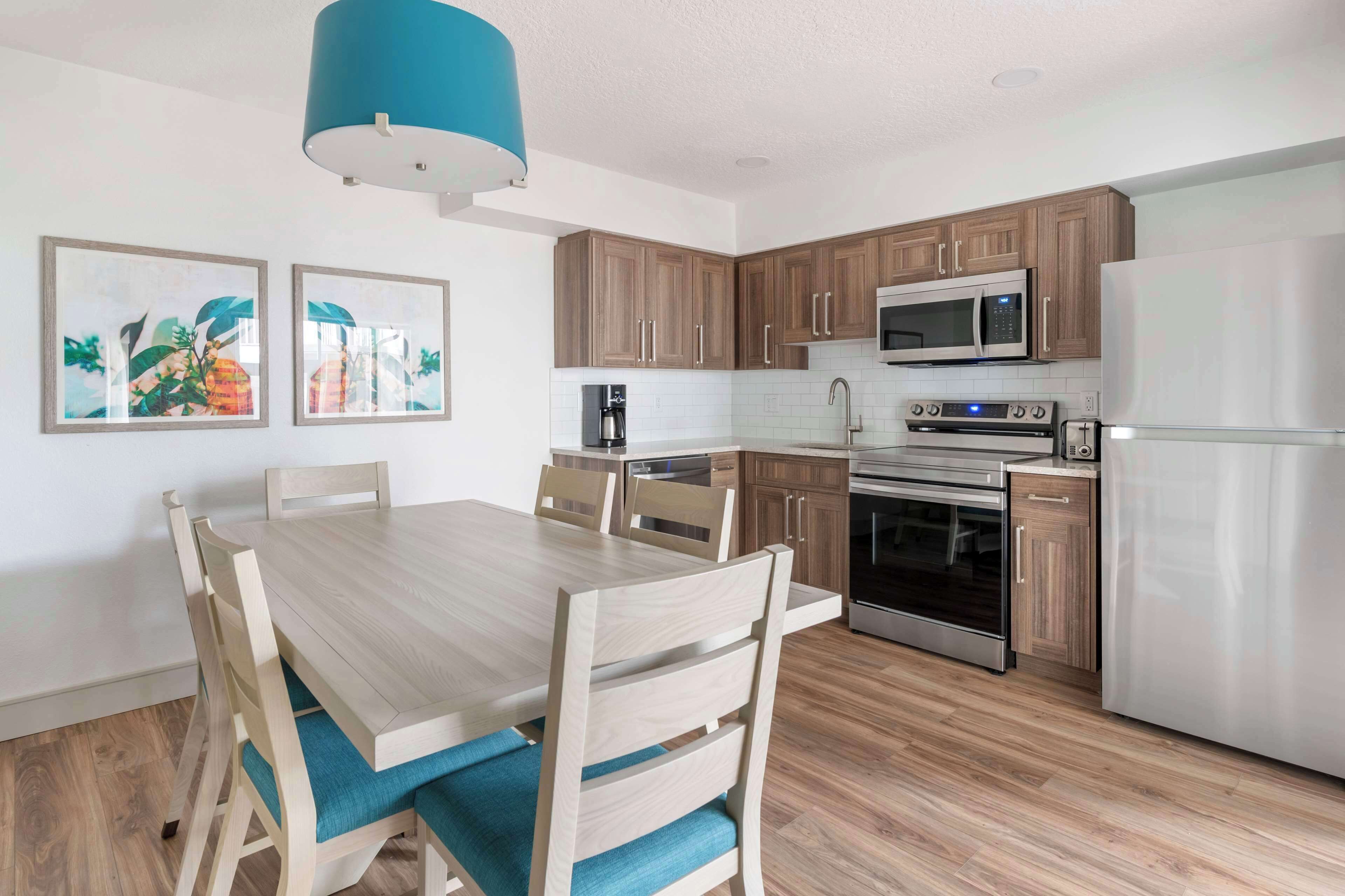 A modern kitchen with wooden cabinets, a stainless steel refrigerator, a microwave, and an oven. There is a dining table with chairs and a blue cushion.