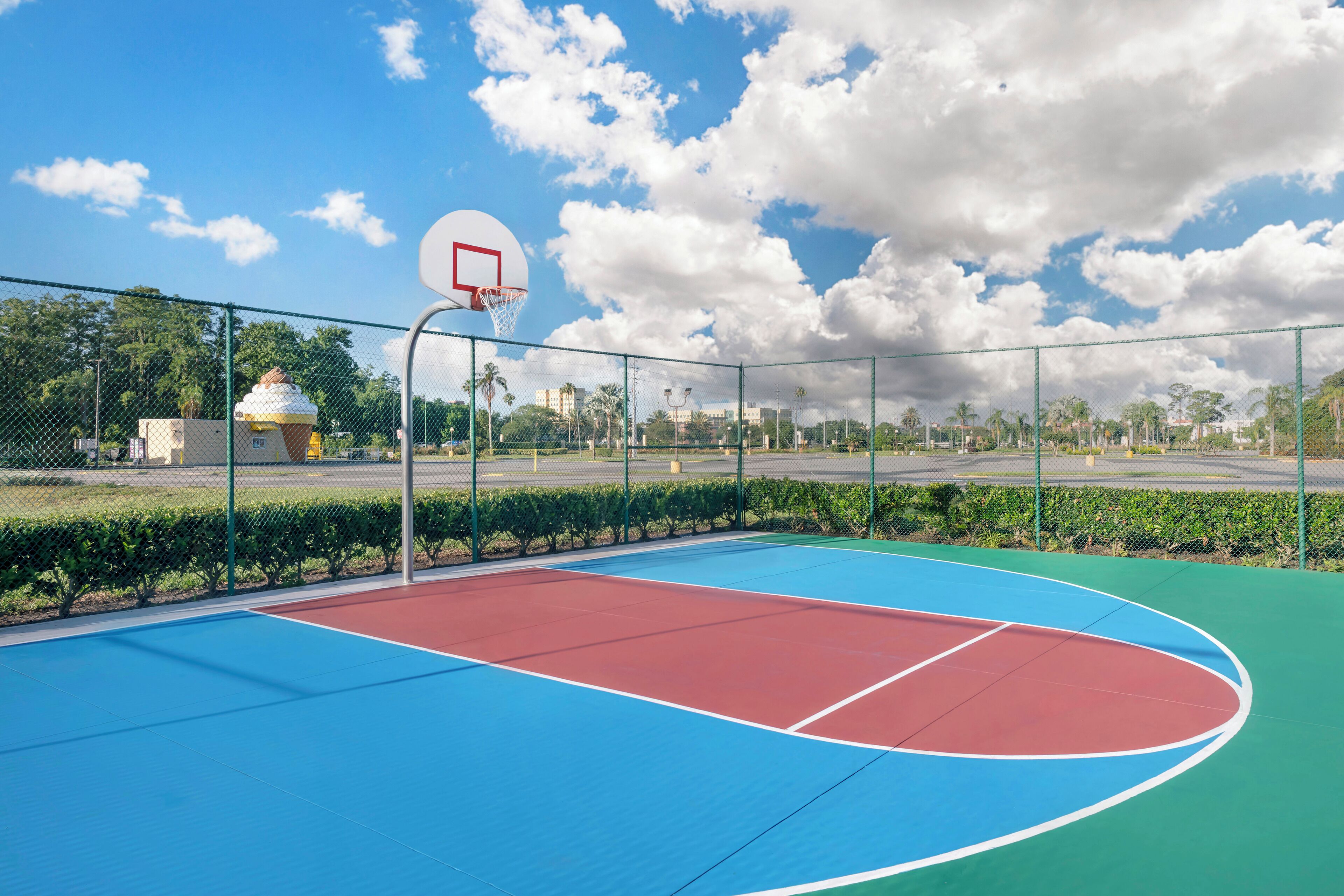 An outdoor basketball court with a hoop, surrounded by a chain-link fence and greenery.
