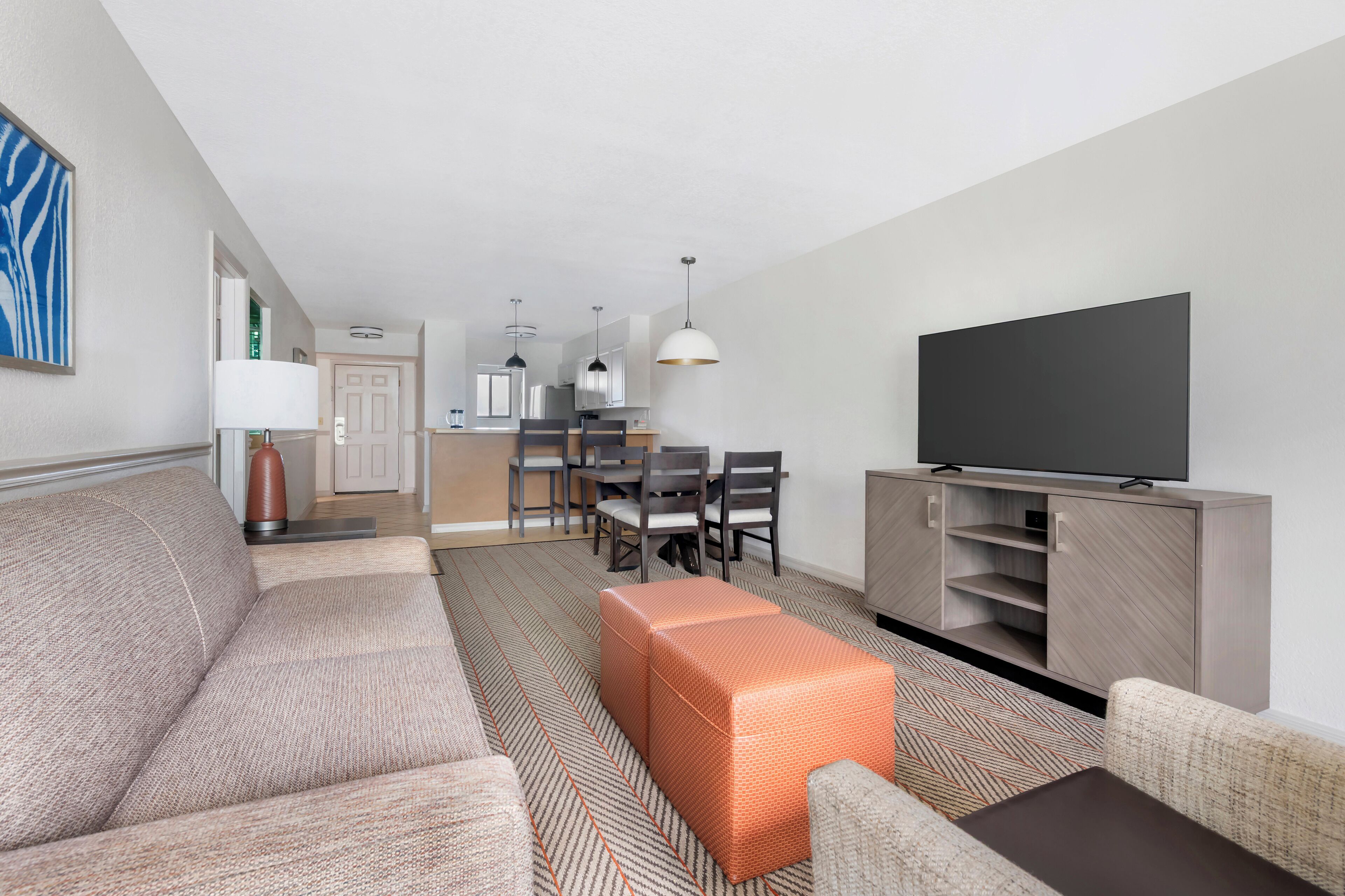 Una sala moderna con un sofá, un reposapiés y un televisor de pantalla plana sobre un mueble de madera. Se puede ver en el fondo un comedor con sillas y una mesa.