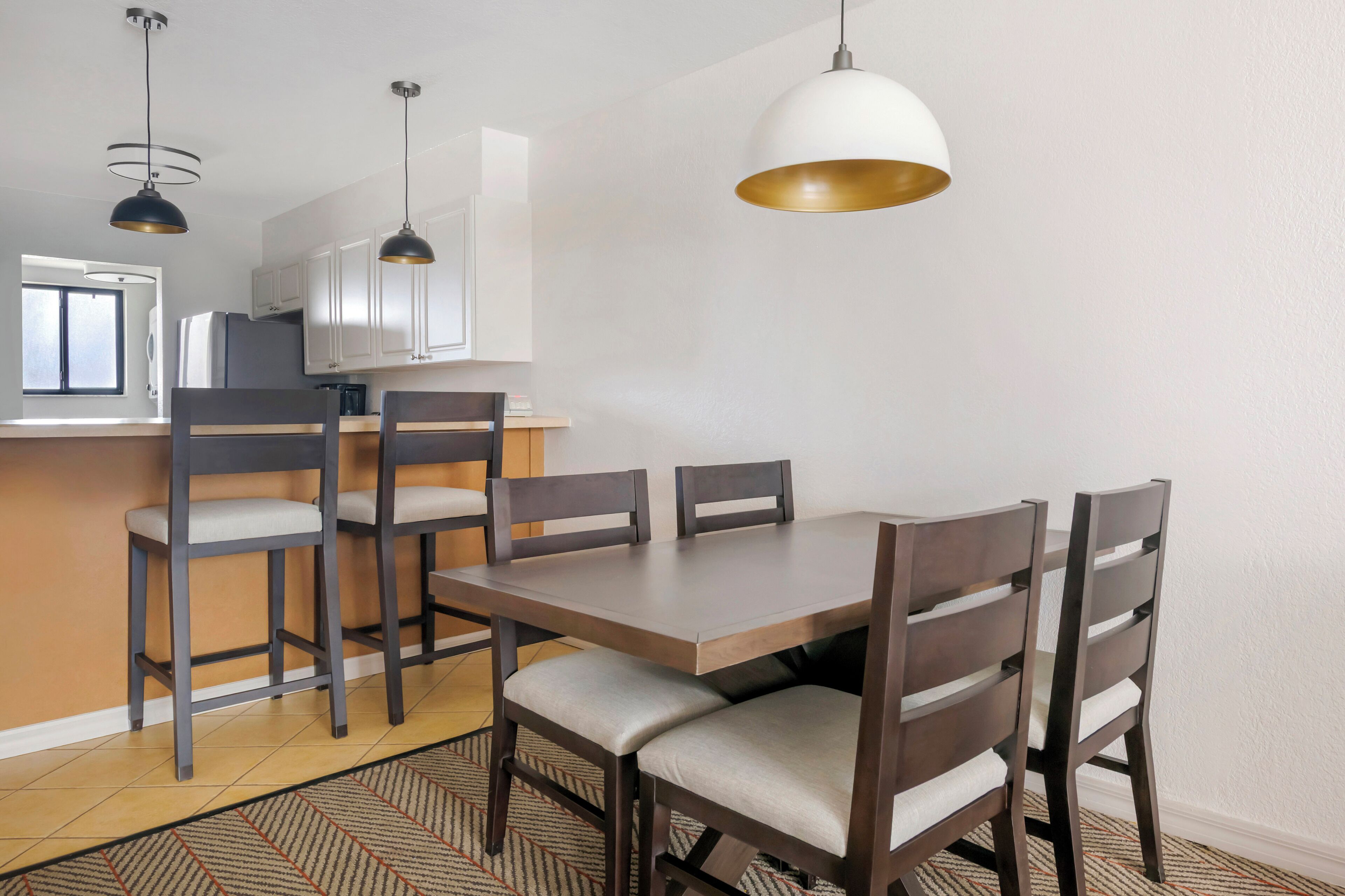 Uma área de refeições com mesa e cadeiras de madeira, cozinha ao fundo e iluminação pendente.