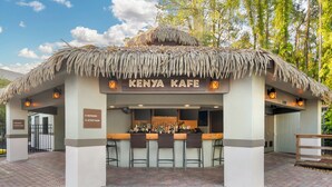 Un bar con techo de paja llamado "Kenya Kafe", con barra, taburetes y un cartel que indica los baños y una sala de actividades.
