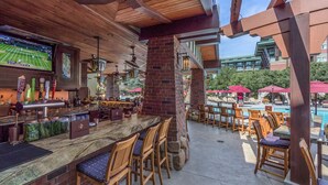 A bar area with a poolside seating arrangement, a bar counter, and a television screen displaying a tennis match.