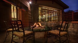Un área de comedor al aire libre con un pozo de fuego rodeado de sillas, un muro de piedra y un edificio con grandes ventanales al fondo.