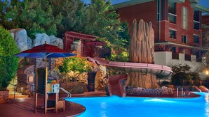 A pool area with a waterfall feature, a wooden deck, and a red building in the background.