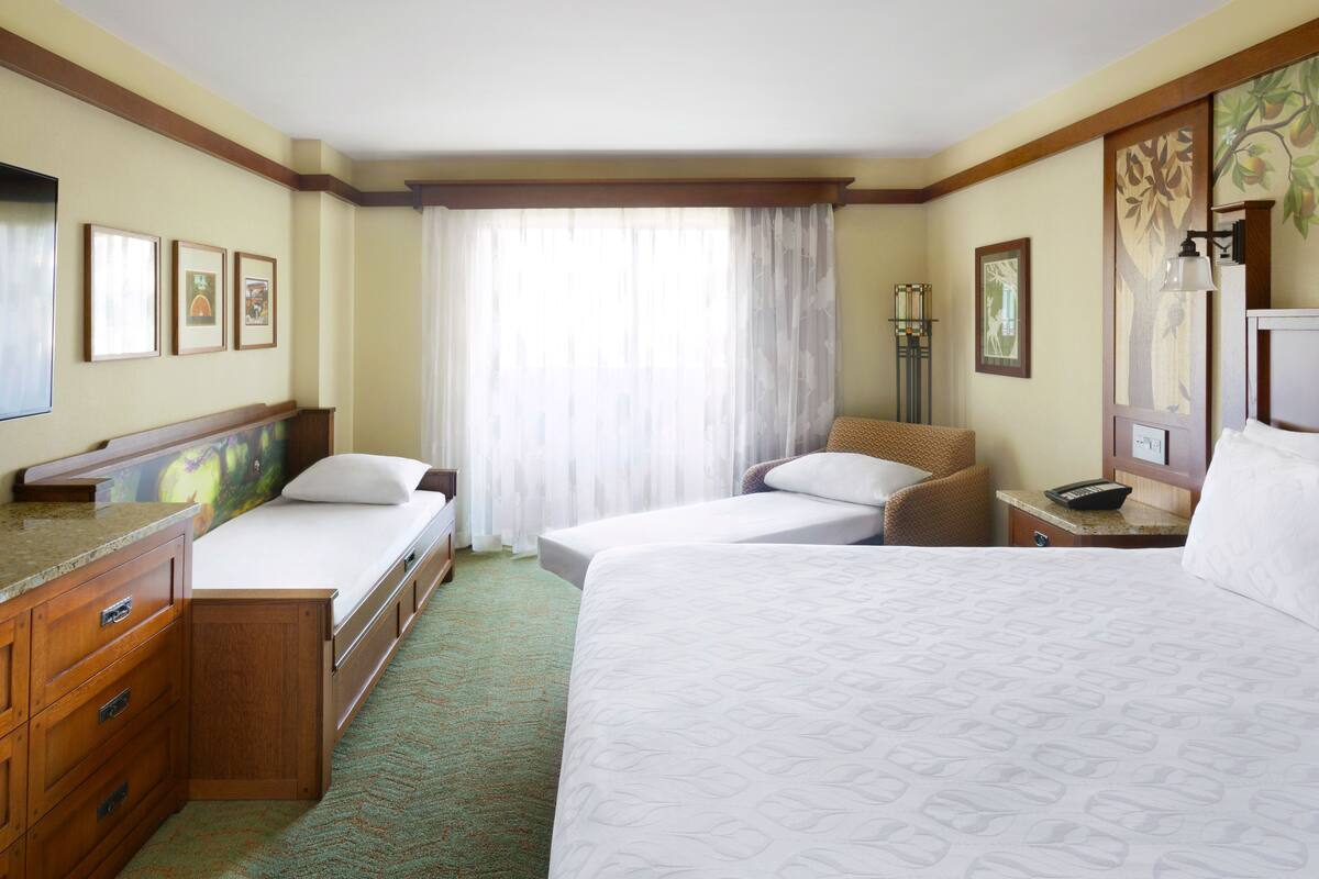 Een hotelkamer met twee eenpersoonsbedden, een houten dressoir, een bureau met lamp en een groot raam met dunne gordijnen.