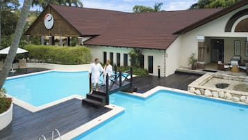 A swimming pool with a wooden deck, a building with a clock, and palm trees in the background.
