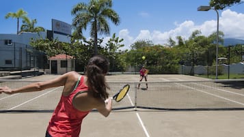 Una cancha de tenis con red, palmeras y un edificio con un letrero al fondo.
