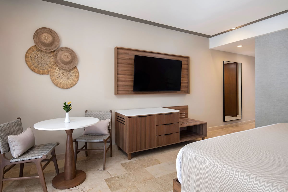 Une chambre d’hôtel moderne comprenant un lit, un téléviseur fixé au mur, une armoire en bois, une petite table ronde avec un vase et deux chaises.