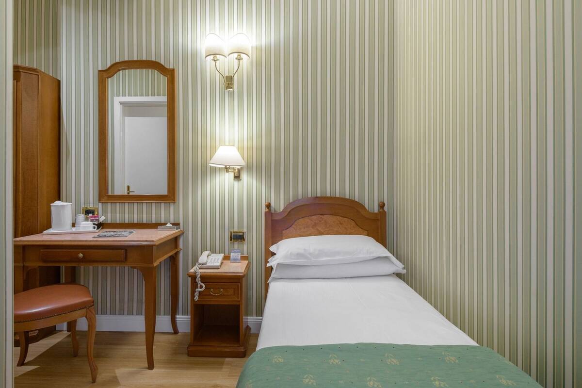 Une chambre d’hôtel avec un bureau en bois, une chaise et un lit recouvert d’une couverture verte. Un miroir est placé au-dessus du bureau et des lampes sont fixées au mur.