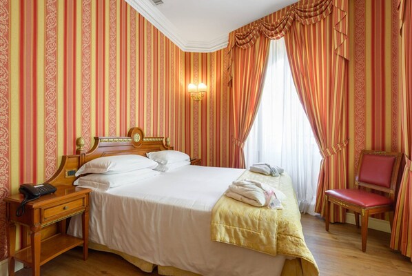 A hotel room with a large bed, a wooden nightstand with a telephone, a red chair, and a window with curtains.