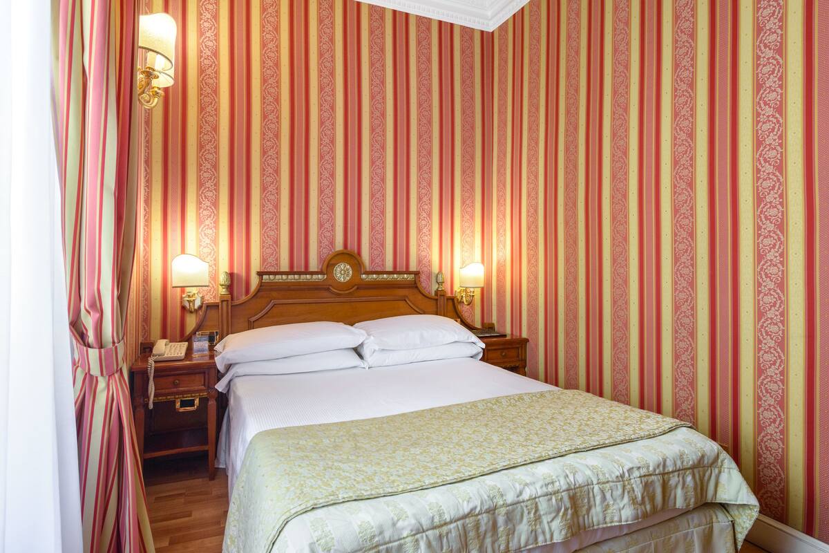 Une chambre d’hôtel avec un grand lit, deux tables de chevet avec des lampes, une tête de lit en bois et un papier peint à rayures.