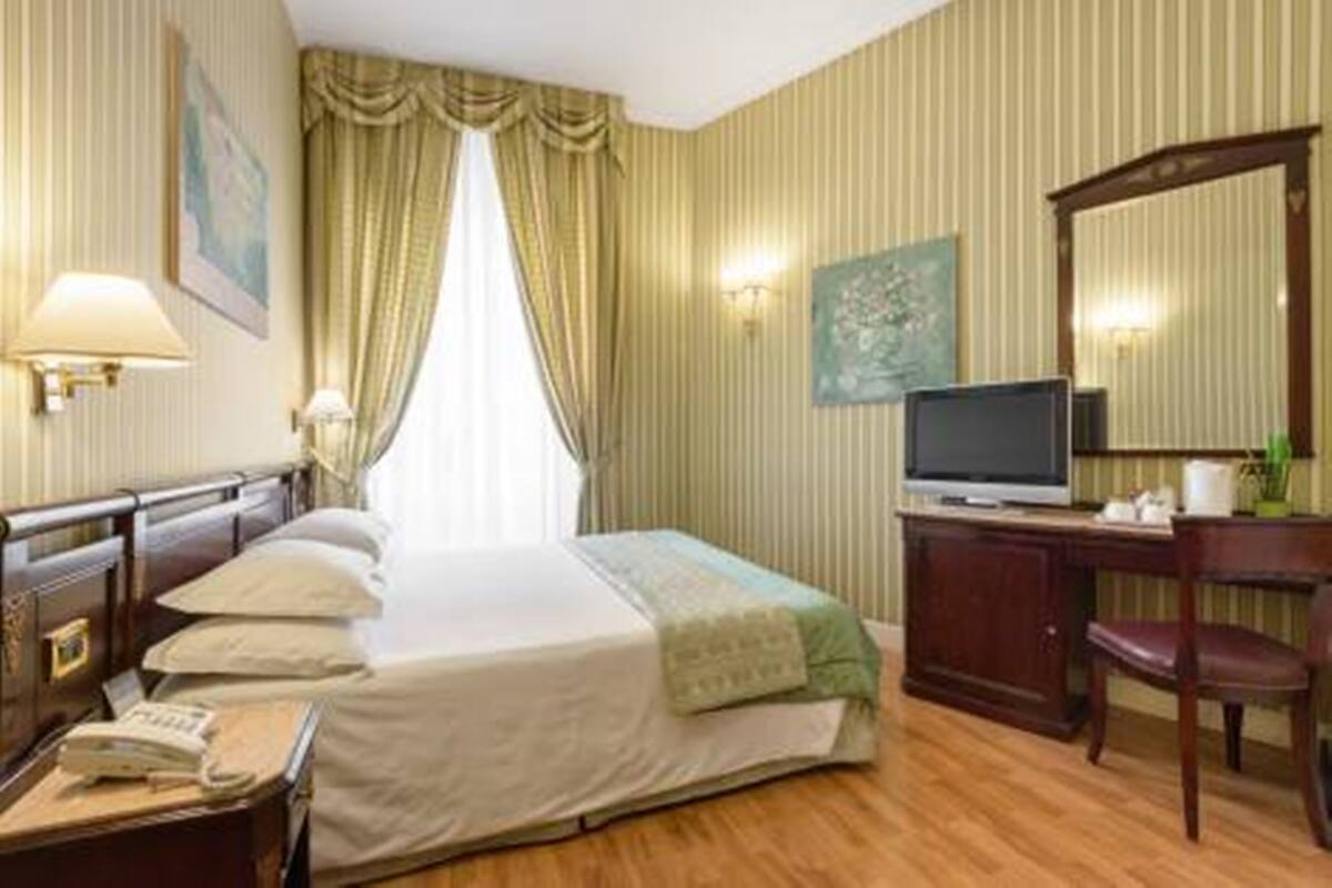 Une chambre d’hôtel comprenant un lit, une table de chevet, une lampe, une télévision et un bureau avec une chaise.