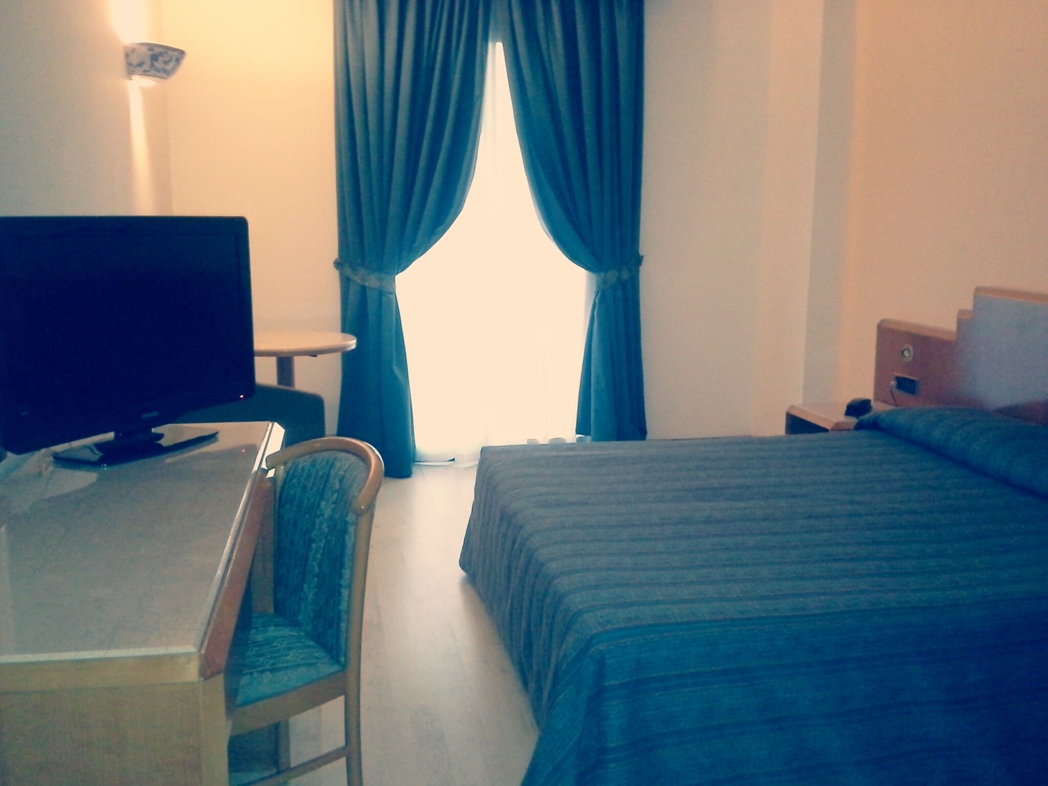 Una camera d'albergo con un letto, una scrivania con una sedia, una televisione e delle tende.