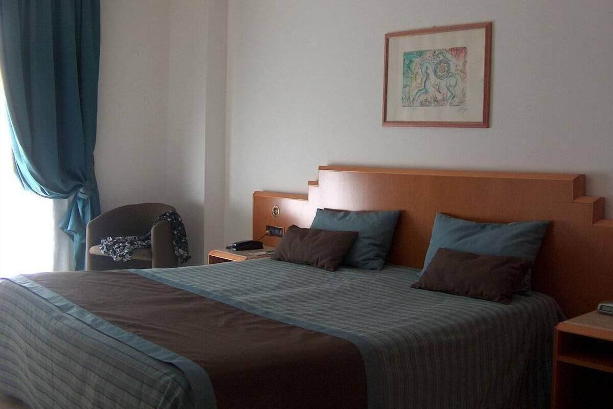 A hotel room with a large bed, a wooden headboard, a nightstand, a chair, and a painting on the wall.