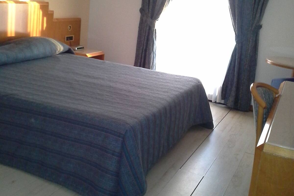 A hotel room with a large bed, a nightstand, a chair, and a window with curtains.