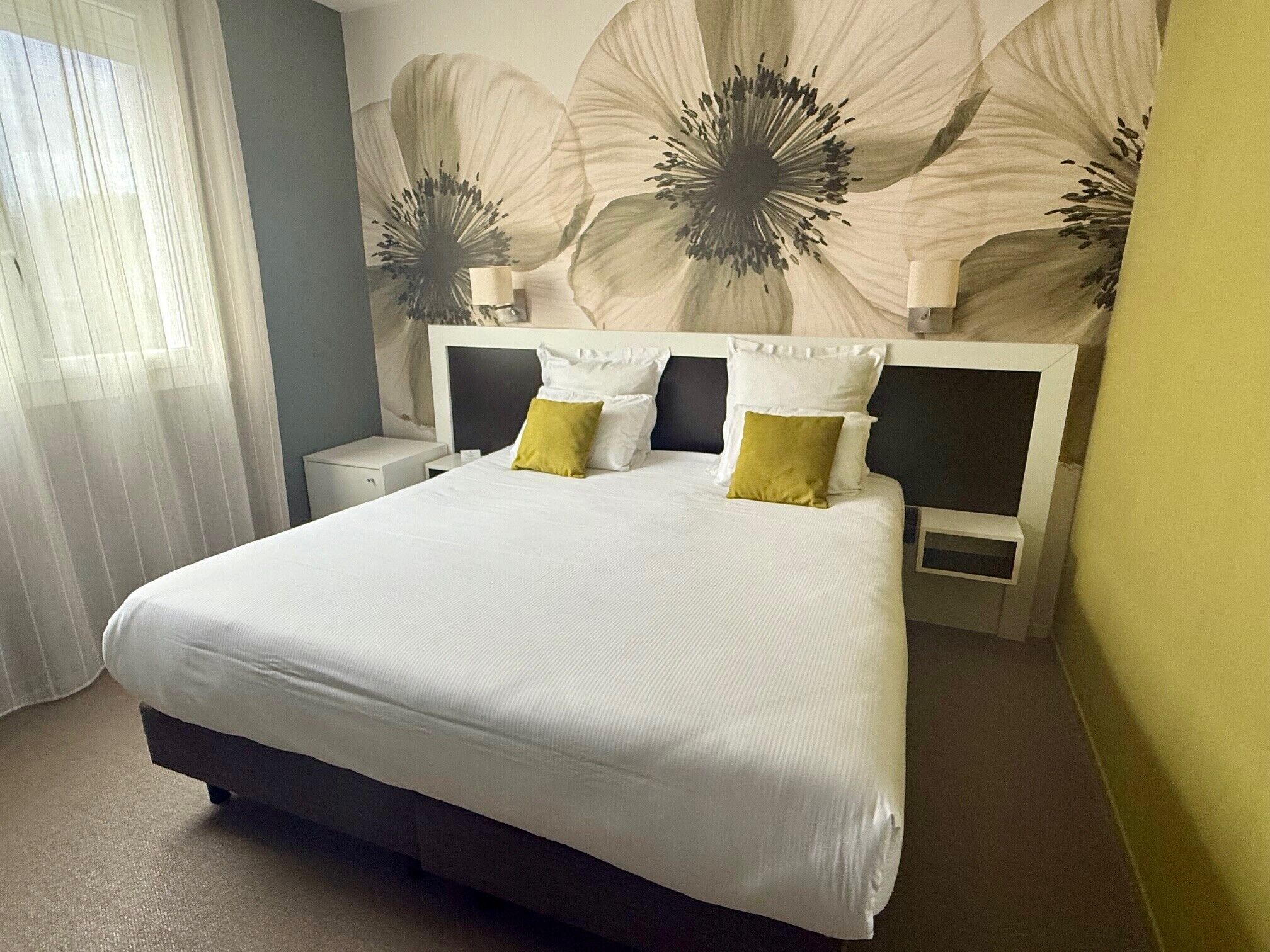 Una cama bien tendida con almohadas blancas y amarillas, un cabecero oscuro y una pared con estampado floral grande.