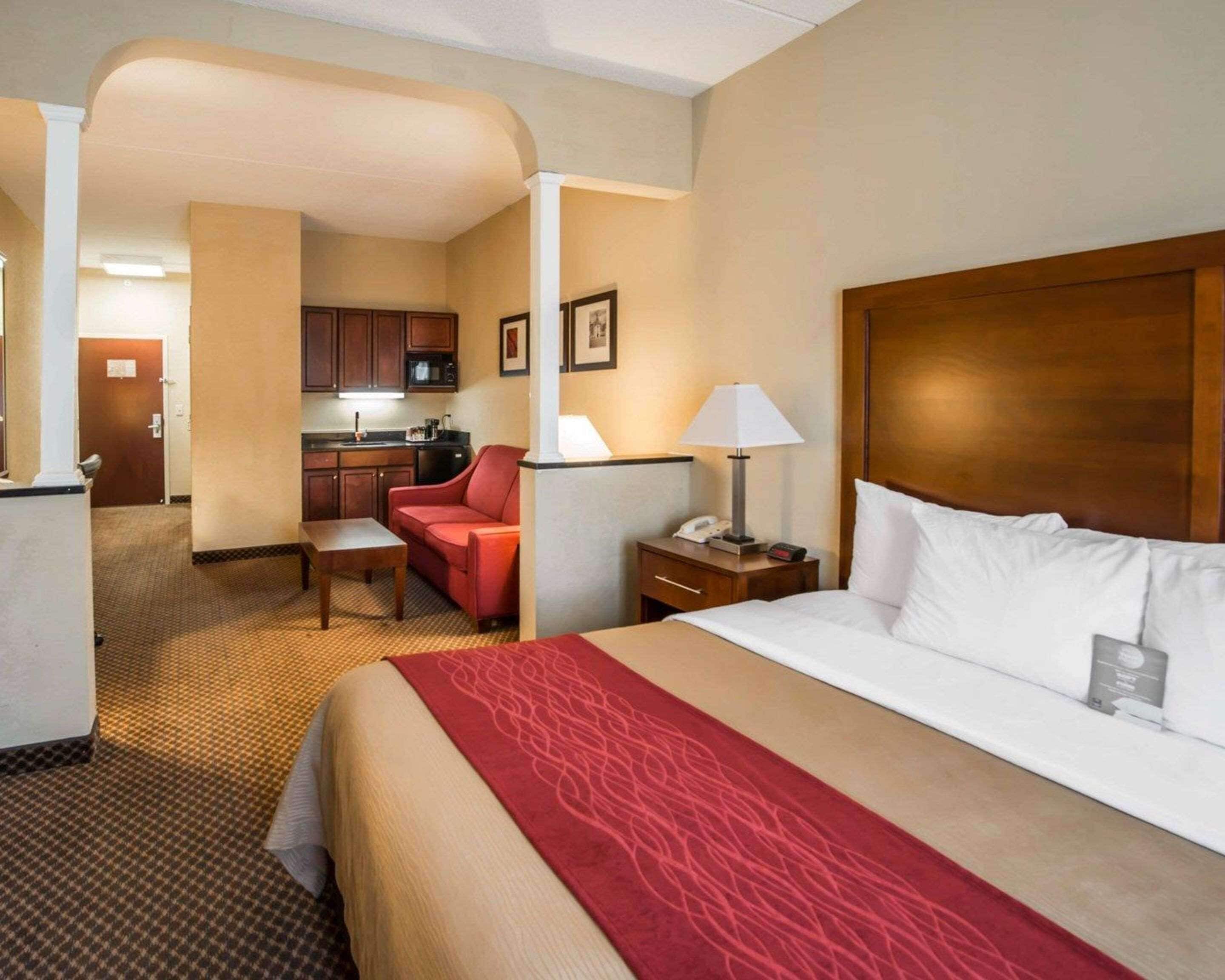 A hotel room with a large bed, a red sofa, a small table, a nightstand with a lamp, a kitchenette, and a door leading to another room.