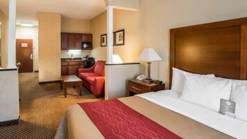 A hotel room with a large bed, a red sofa, a small table, a nightstand with a lamp, a kitchenette, and a door leading to another room.