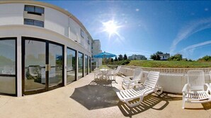 Um edifício moderno com fachada curva, grandes portas de vidro e um espaço de varanda com cadeiras de lounge brancas e um guarda-sol azul.