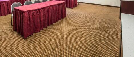 Uma sala de conferências com mesas compridas cobertas por toalhas vermelhas, uma tela de projetor e piso carpeteado.