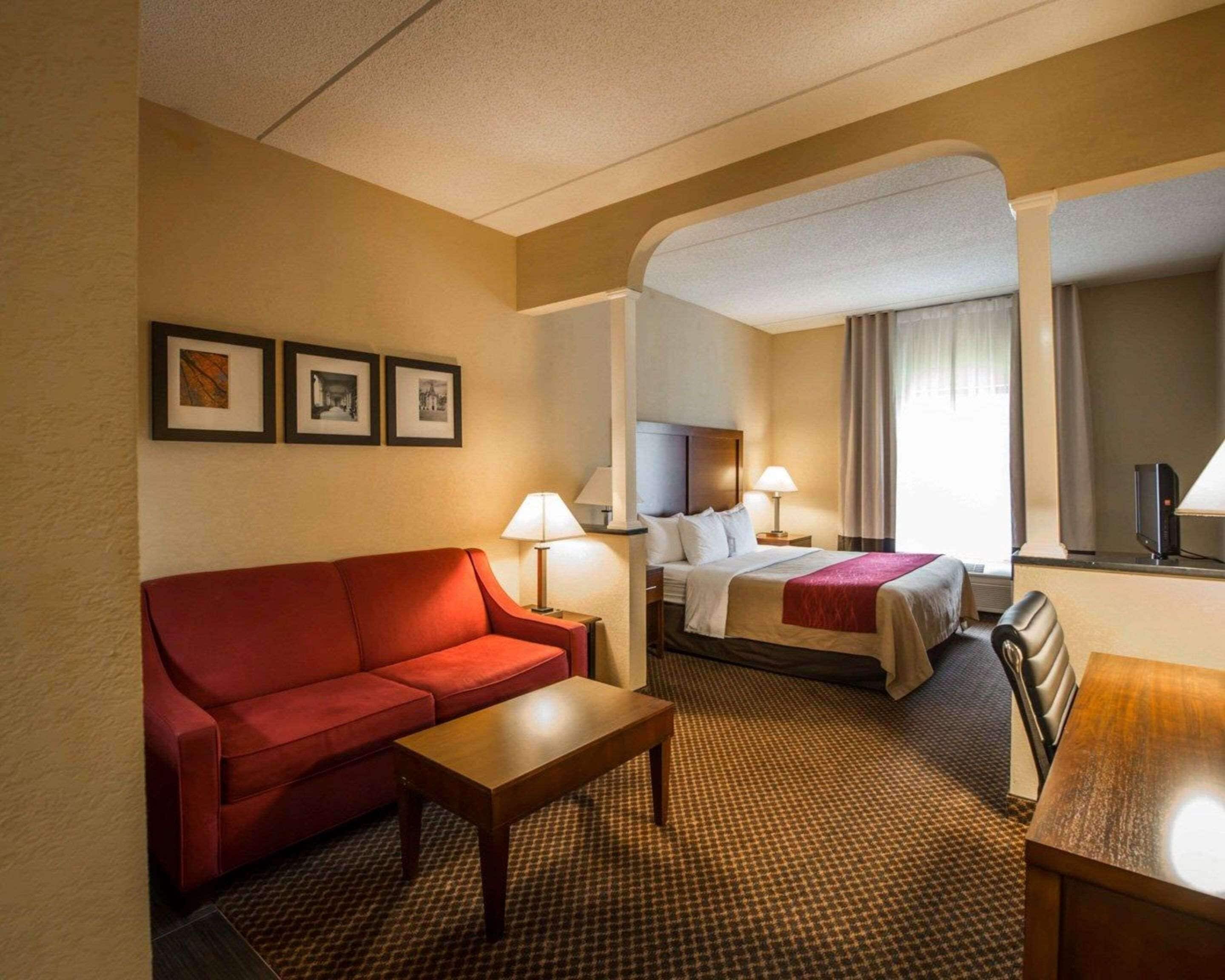 Une chambre d’hôtel avec un canapé rouge, une table basse en bois, un lit recouvert d’une couette blanche et un bureau avec une lampe.