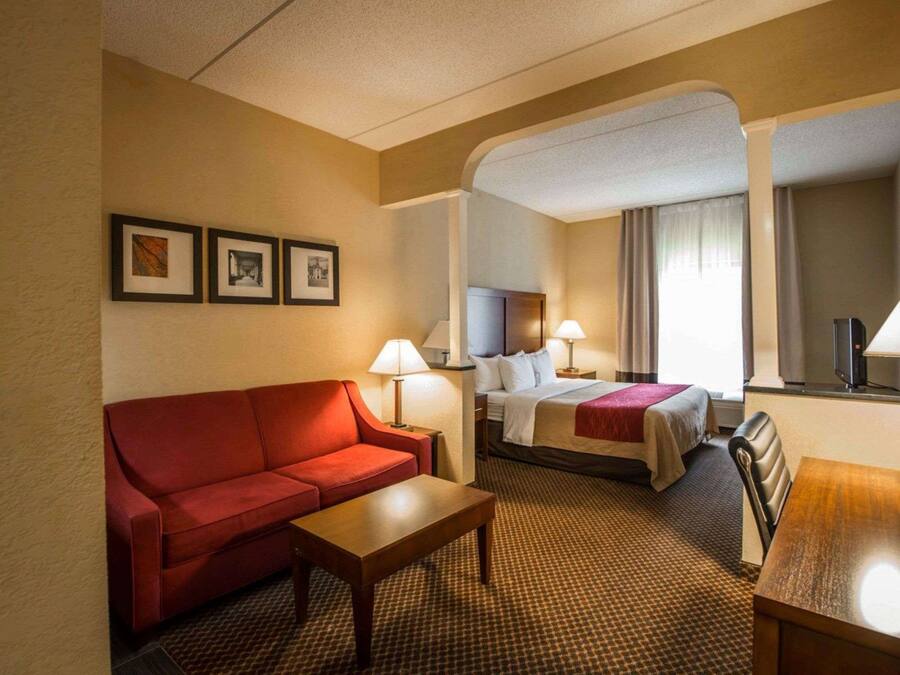 Une chambre d’hôtel avec un canapé rouge, une table basse en bois, un lit recouvert d’une couette blanche et un bureau avec une lampe.