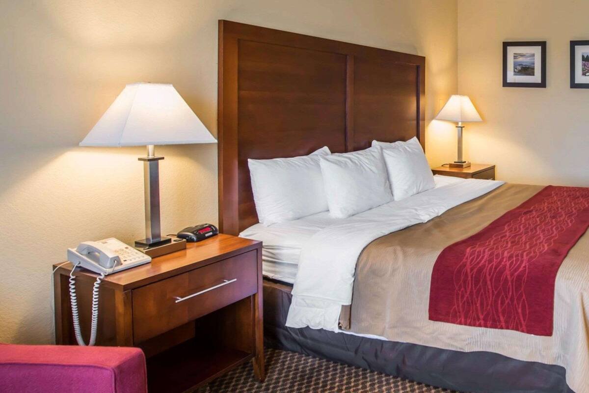 A hotel room with a wooden headboard, a bed with white linens, a red and beige throw blanket, a desk with a telephone and a lamp, and a patterned carpet.