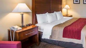 A hotel room with a wooden headboard, a bed with white linens, a red and beige throw blanket, a desk with a telephone and a lamp, and a patterned carpet.