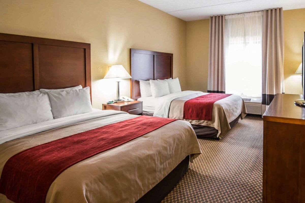 A hotel room with two beds, a nightstand with a lamp, a desk, and a window with curtains.