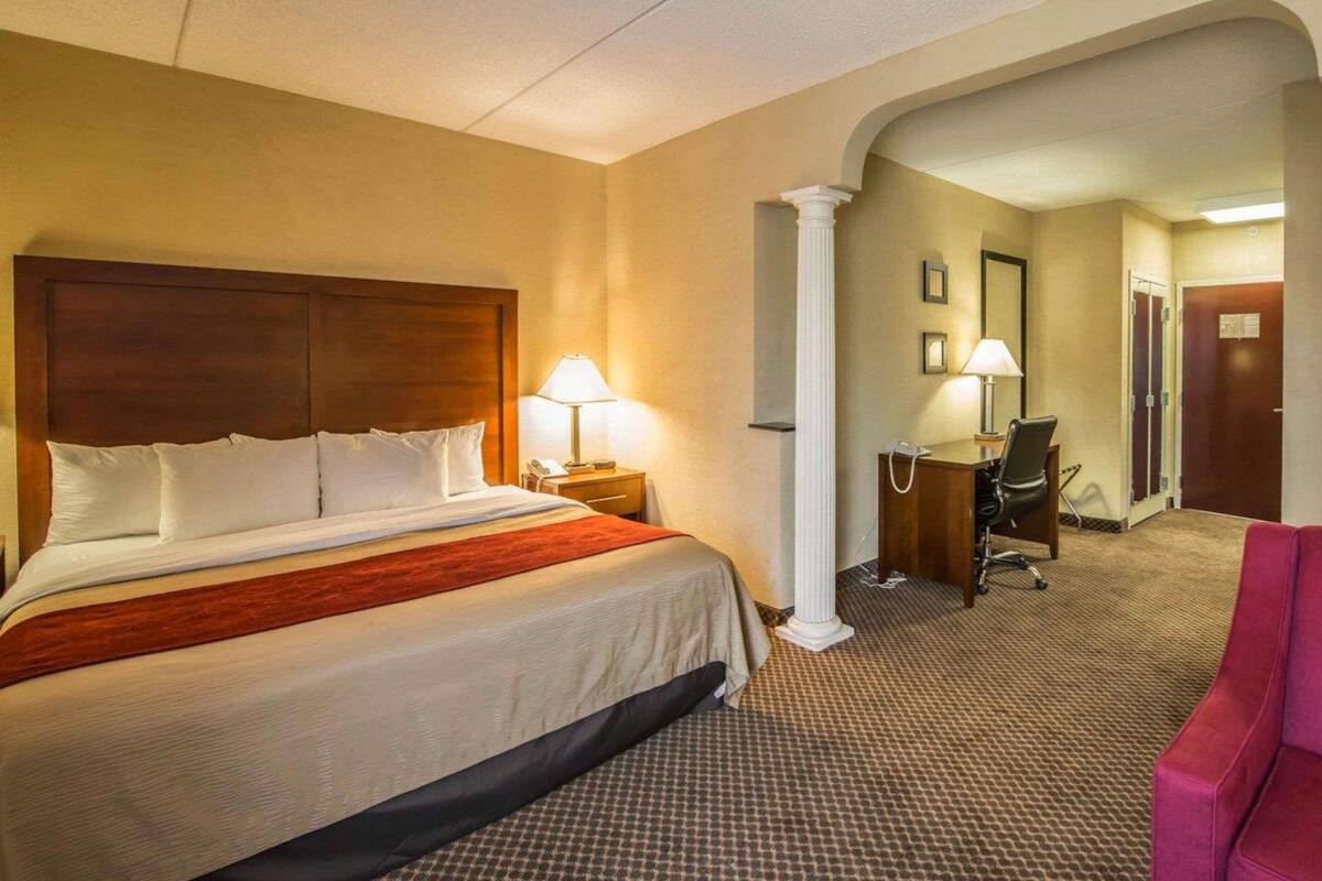 A hotel room with a large bed, two bedside lamps, a desk with a chair, and a pink armchair.